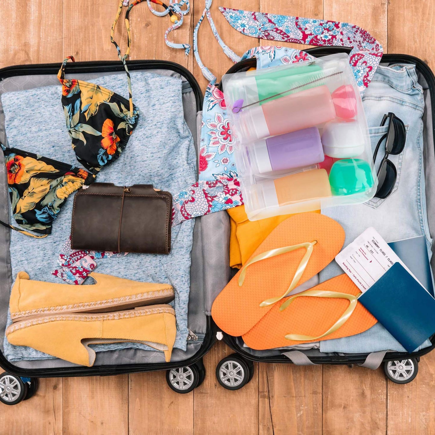 A packed suitcase with colorful swimsuits, a wallet, toiletries, flip-flops, and clothing, set against a wooden floor.